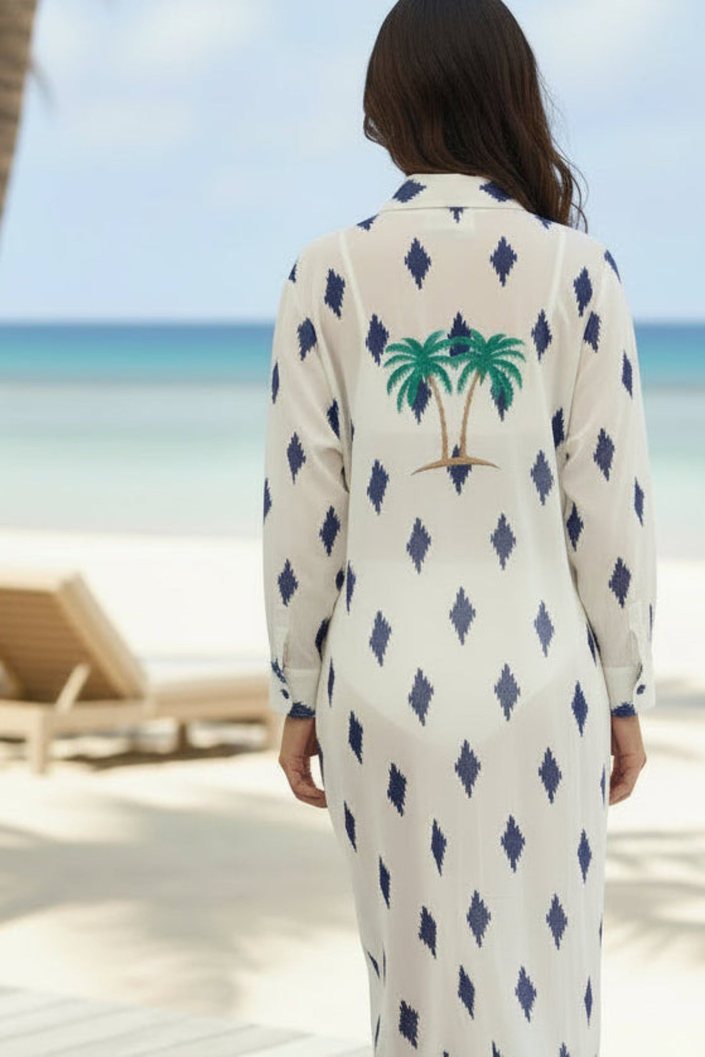 Woman in a patterned dress walking on a wooden deck with a beach and palm tree in the background